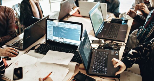 Profissionais utilizam computadores e notebooks em ambiente corporativo. A imagem ilustra discussões sobre o fim da escala 6x1, jornada de trabalho de 32 horas, inteligência artificial e produtividade.