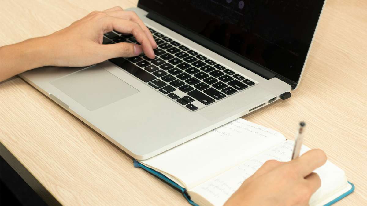 pessoa digitando em um notebook com um caderno ao lado