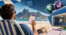 Homem de costas segura um volante da Lotofácil em uma praia, com uma casa lotérica e bolas coloridas ao fundo.
