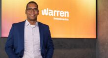Homem de óculos e terno azul posando em frente a um painel com o logo Warren Investimentos.