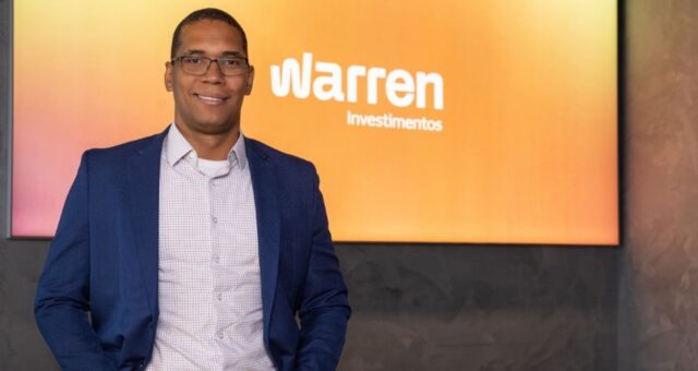 Homem de óculos e terno azul posando em frente a um painel com o logo Warren Investimentos.