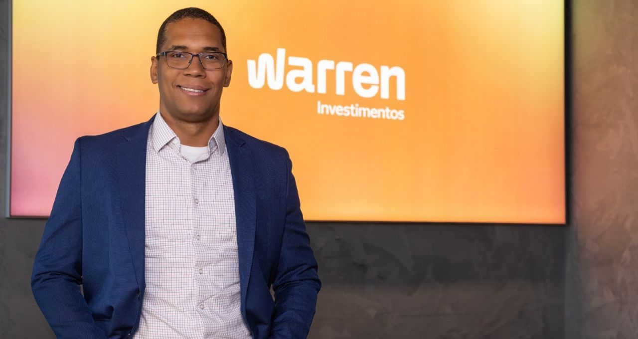 Homem de óculos e terno azul posando em frente a um painel com o logo Warren Investimentos.